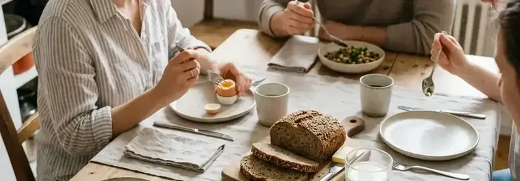 Ein gedeckter Frühstückstisch aus hellem Holz mit Skyr-Beeren-Schale, gekochten Eiern und Brot von oben im Morgenlicht
